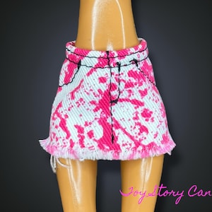 Puede incluir: Minifalda de mezclilla con una base azul claro y un vibrante estampado de salpicaduras rosas. La falda presenta costuras negras y un dobladillo deshilachado, diseñada para una muñeca.