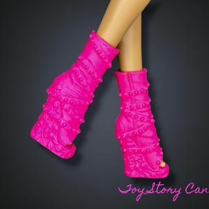 Peut inclure: Bottes de poupée rose vif avec un design texturé et des clous. Les bottes à bout ouvert sont sur un fond gris foncé. Les jambes de la poupée sont visibles.