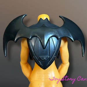 Puede incluir: Accesorio de ala de murciélago de plástico negro adjunto a una figura de acción amarilla. Las alas de murciélago tienen bordes curvos y una pieza central en forma de escudo. La figura tiene cabeza y torso amarillos.