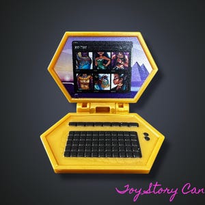 Op de afbeelding: Een gele speelgoedlaptop met een zwart toetsenbord en scherm met afbeeldingen van vrouwen. Het scherm wordt omlijst door een paarse rand met een piramide- en zonsondergangachtergrond. Het speelgoed is open en toont het scherm en het toetsenbord.