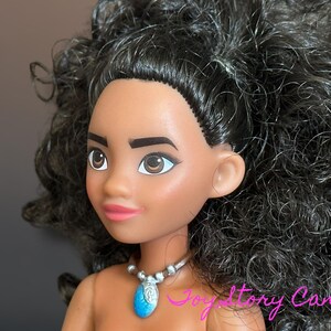 May include: A doll with dark skin, brown eyes, and black curly hair. The doll is wearing a silver necklace with a blue pendant. The doll's lips are painted pink. The doll is a character from a popular animated movie.