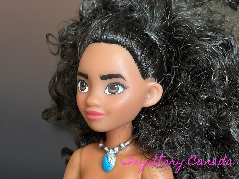 May include: A doll with dark skin, brown eyes, and black curly hair. The doll is wearing a silver necklace with a blue pendant. The doll's lips are painted pink. The doll is a character from a popular animated movie.