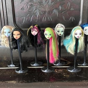 Könnte beinhalten: Sammlung von Puppenköpfen mit verschiedenen Frisuren und Make-up. Die Köpfe sind auf schwarzen Ständern montiert. Die Haarfarben umfassen Blond, Schwarz, Pink, Grün und Blau. Die Puppen haben detaillierte Gesichtszüge.