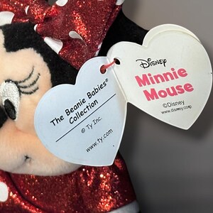 May include: A Minnie Mouse plush toy from The Beanie Babies Collection. The toy features a red sequined dress and bow with white polka dots. The heart-shaped tags read "Disney Minnie Mouse" and "The Beanie Babies Collection."