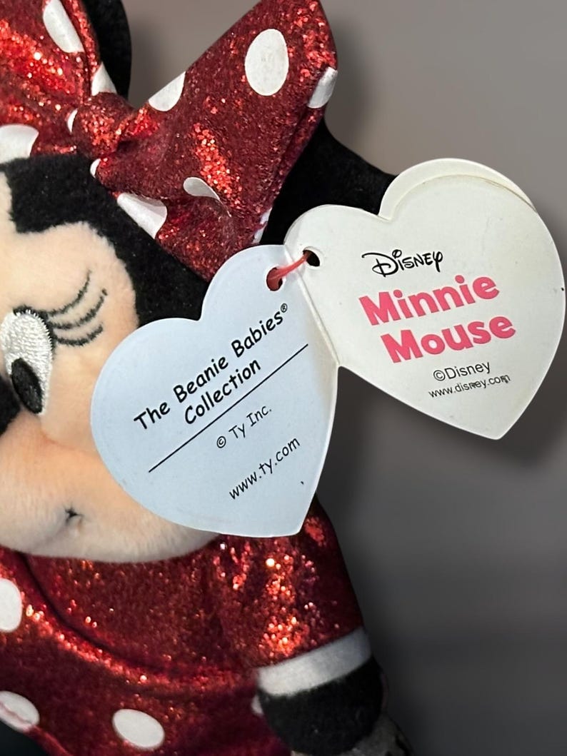 May include: A Minnie Mouse plush toy from The Beanie Babies Collection. The toy features a red sequined dress and bow with white polka dots. The heart-shaped tags read "Disney Minnie Mouse" and "The Beanie Babies Collection."