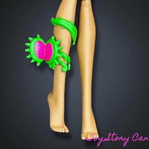 Puede incluir: Piernas de muñeca con un accesorio verde y rosa con temática de Venus atrapamoscas. El accesorio está unido a la pierna de la muñeca. Las piernas son de color bronceado claro y el fondo es gris oscuro.