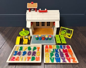 May include: A vintage Fisher-Price Little People Schoolhouse playset with a white building, a green playground, and a set of colorful alphabet and number blocks. The schoolhouse has a red roof and a bell on top. The playground includes a slide, a swing set, and a spinning wheel. The alphabet and number blocks are in a white tray.