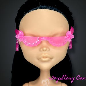 Puede incluir: Una cabeza de muñeca con gafas de sol rosas con flores en las sienes. Las gafas tienen un borde ondulado. La muñeca tiene el pelo negro y tez clara. El fondo es oscuro.
