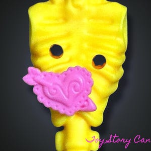 Puede incluir: Un objeto amarillo, estilizado en forma de torso, con dos agujeros circulares y un adorno en forma de corazón rosa. El objeto tiene una superficie texturizada y está sobre un fondo oscuro. El texto "JoyStory Canada" es visible.