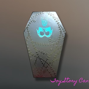 Puede incluir: Una caja plateada en forma de ataúd con un diseño cosido y un gráfico de máscara turquesa. La caja tiene el texto "Monster High" en la parte superior. El diseño incluye imperdibles y un patrón cosido.