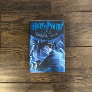 May include: A blue and white book cover with the title "Harry Potter and the Order of the Phoenix" and the author's name "J.K. Rowling". The cover features a young boy with dark hair and glasses, wearing a dark robe, looking to the right.