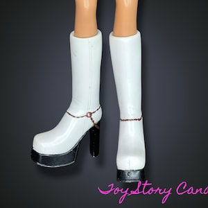 Puede incluir: Botas blancas de plataforma con tacones negros y cadenas en el tobillo. Las botas son hasta la rodilla y tienen un acabado brillante. La imagen está sobre un fondo oscuro. Las botas miden unos 25 cm de alto.