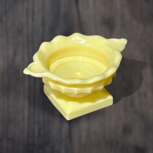 Könnte beinhalten: Ein gelber Keramiktopf mit einem gewellten Rand und einem quadratischen Sockel. Der Topf ist für eine kleine Pflanze oder Blume gedacht.