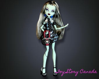 May include: A Monster High doll with green skin, long gray hair, and a black and white plaid dress. The doll is holding a red and black purse with a skull design and wearing black and white striped heels.