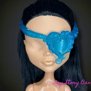 Könnte beinhalten: Eine Puppe mit schwarzen Haaren und hellem Hautton trägt eine durchscheinende blaue Augenklappe. Die Augenklappe ist herzförmig mit dekorativen Wirbeln und einem Band, das um den Kopf geht.