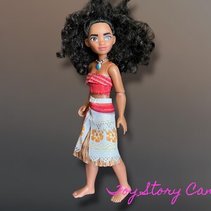 May include: A doll of a Polynesian woman with black curly hair, wearing a red top, a white skirt with orange floral patterns, and a blue necklace. The doll is standing on a brown surface.