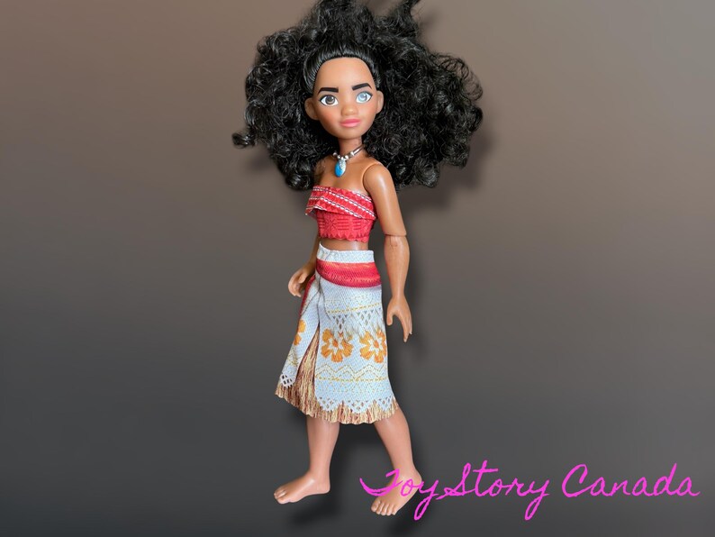 May include: A doll of a Polynesian woman with black curly hair, wearing a red top, a white skirt with orange floral patterns, and a blue necklace. The doll is standing on a brown surface.