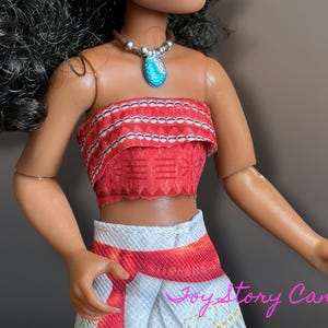May include: A doll wearing a red and white patterned crop top and skirt. The doll has a silver necklace with a blue pendant. The doll's skin is tan, and it has black hair. The doll is wearing a brown necklace.