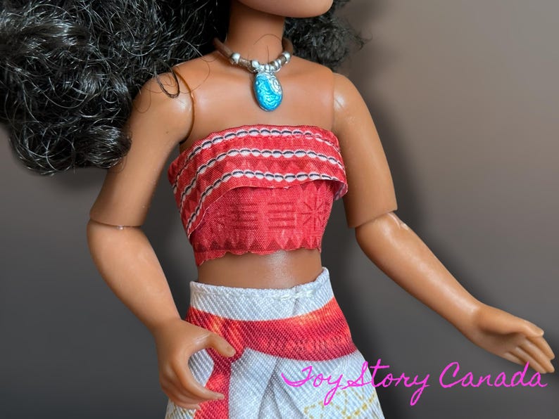 May include: A doll wearing a red and white patterned crop top and skirt. The doll has a silver necklace with a blue pendant. The doll's skin is tan, and it has black hair. The doll is wearing a brown necklace.