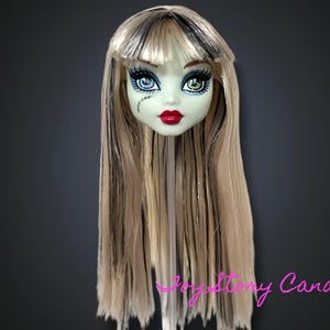 Puede incluir: Cabeza de muñeca con piel verde pálido, labios rojos y ojos desiguales. La muñeca tiene pelo largo, castaño claro con mechones negros y flequillo negro. Una cicatriz cosida es visible en la cara de la muñeca.