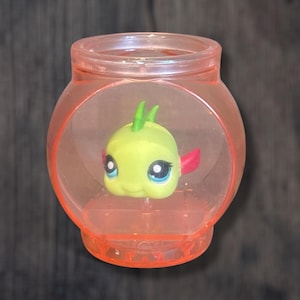 May include: A clear plastic fishbowl with a green and pink fish figurine inside. The fishbowl is round and has a light orange tint.