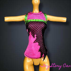 Puede incluir: Una camiseta para muñeca en rosa brillante con una superposición de red negra. La camiseta tiene ribetes verdes alrededor del cuello y los hombros. La muñeca lleva ropa interior amarilla con lunares. El fondo es gris oscuro.