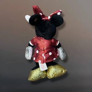 May include: A plush Minnie Mouse doll, viewed from the back, features a red dress with white polka dots, a matching bow, and gold glitter shoes. The doll has black ears, a black head, and silver glitter gloves.