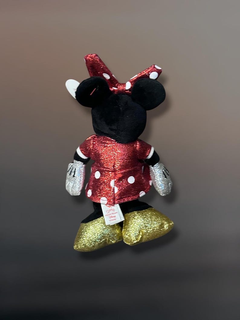 May include: A plush Minnie Mouse doll, viewed from the back, features a red dress with white polka dots, a matching bow, and gold glitter shoes. The doll has black ears, a black head, and silver glitter gloves.