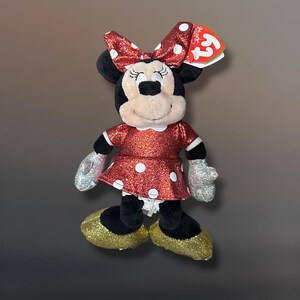 May include: A plush Minnie Mouse doll with a red glittery dress and bow, both adorned with white polka dots. The doll has black ears, a tan face, silver gloves, and gold glitter shoes. A Ty tag is attached.