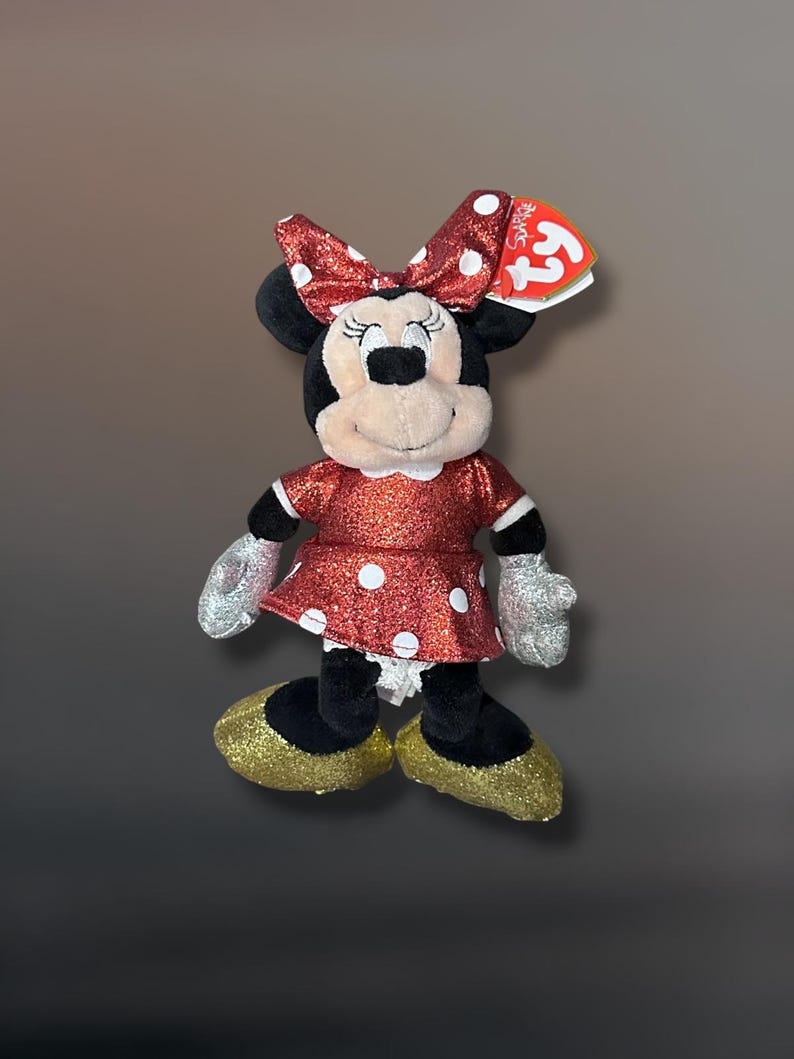 May include: A plush Minnie Mouse doll with a red glittery dress and bow, both adorned with white polka dots. The doll has black ears, a tan face, silver gloves, and gold glitter shoes. A Ty tag is attached.