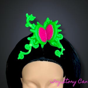 Puede incluir: Un tocado verde neón y rosa intenso, con forma de flor o planta estilizada, sobre un fondo negro. El tocado se lleva sobre una peluca negra. El diseño presenta un elemento central rosa rodeado de verde.