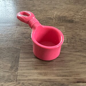May include: A pink plastic measuring cup with a handle. The cup is empty.