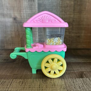 以下が含まれることがあります： ピンクとグリーンのポップコーンカートのおもちゃ。黄色の車輪と、白と黄色のポップコーンが入った透明な容器が付いています。容器には「メインストリートポップコーンカンパニー」の文字が印刷されています。