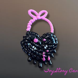 Puede incluir: Colgante negro en forma de corazón con lazo y detalles rosas. El colgante tiene una superficie texturizada con detalles rosas y acentos goteantes. El texto "Joy Story Canada" está en rosa en la parte inferior.