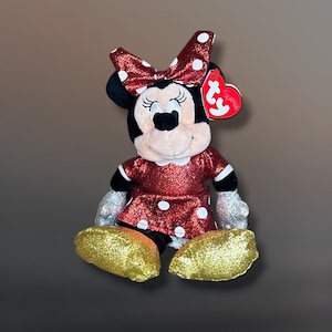 May include: A Minnie Mouse plush toy with a red glitter dress and bow with white polka dots. The plush has gold glitter shoes and silver glitter gloves. A red heart-shaped tag is attached to the ear.