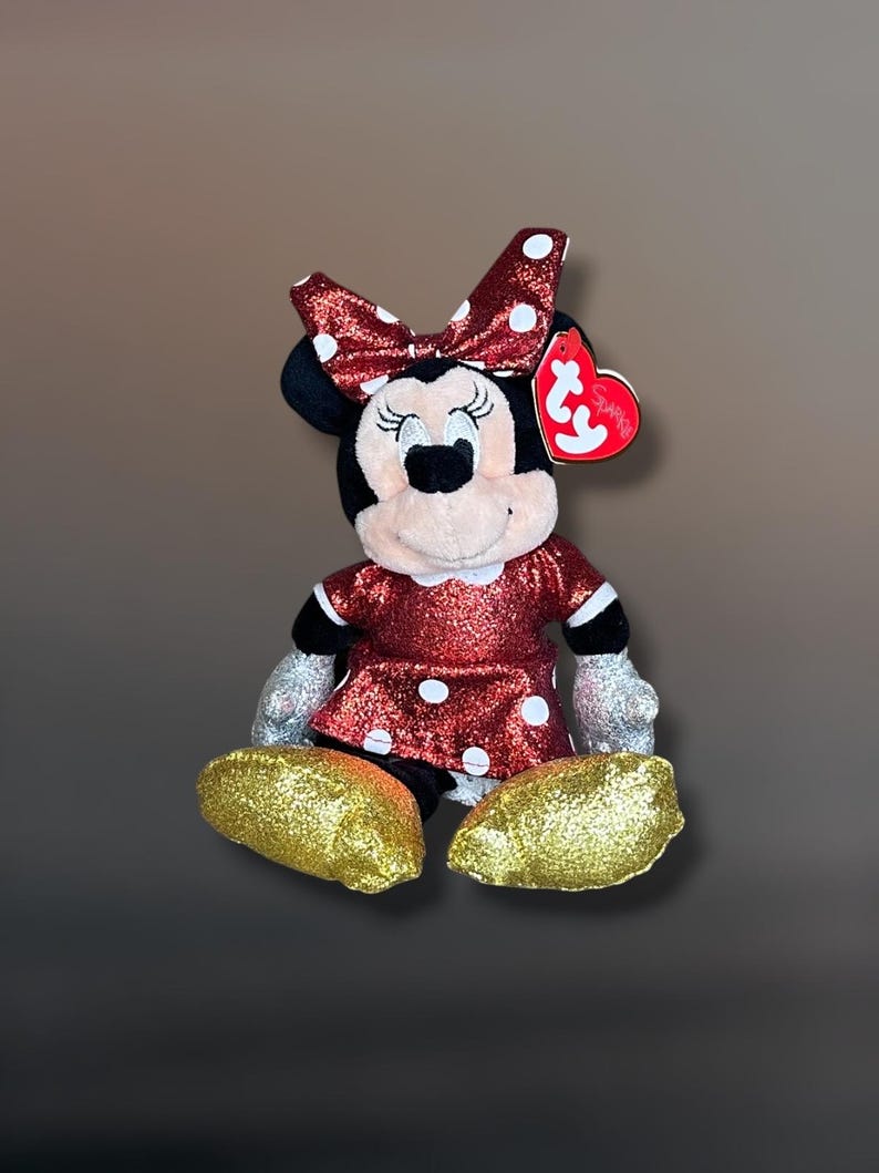 May include: A Minnie Mouse plush toy with a red glitter dress and bow with white polka dots. The plush has gold glitter shoes and silver glitter gloves. A red heart-shaped tag is attached to the ear.