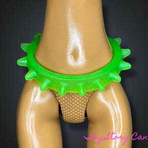 Peut inclure: Un collier à pointes vert fluo. Le collier est fait d'un matériau souple et comporte des pointes pointues espacées uniformément. L'image montre le collier autour du cou d'une poupée, avec un string beige visible en dessous.