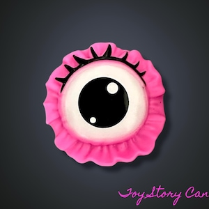 May include: A pink, eye-shaped decorative item with a large black pupil and white iris. The eye is surrounded by a ruffled pink border and black eyelashes. The item is set against a dark grey background.