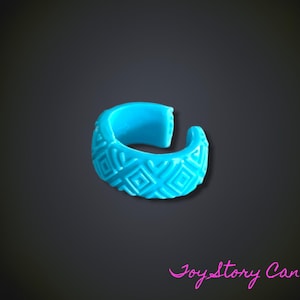 Op de afbeelding: Een fel turquoise ring met een open band en een verhoogd geometrisch patroon van vierkanten en lijnen op de buitenkant. De ring heeft een gladde binnenkant en een opening aan de achterzijde. De tekst Joy Story Canada staat in het roze in de hoek.