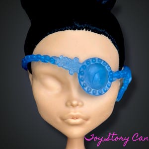 Puede incluir: Una cabeza de muñeca con cabello negro y un parche en el ojo azul. El parche en el ojo es de color azul claro y tiene un diseño circular sobre el ojo. El parche en el ojo está conectado a una banda que rodea la cabeza.