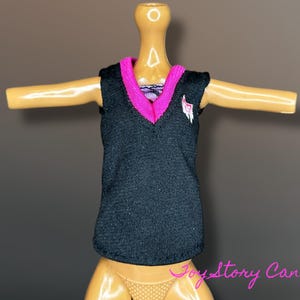 May include: A black doll vest with a pink V-neck collar and trim. A white and pink emblem is on the left chest. The vest is sleeveless and appears to be made of a textured fabric. The doll is posed with arms outstretched.