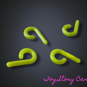 Puede incluir: Cuatro adornos translúcidos en forma de bastón de caramelo verde lima están dispuestos sobre un fondo gris oscuro. Los adornos tienen un gancho curvo y un tallo recto. El texto "Joy Story Canada" está en escritura rosa en la parte inferior.