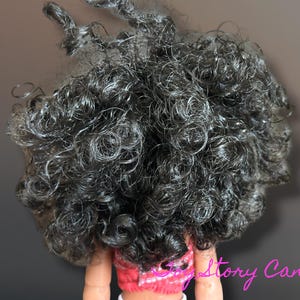 May include: Close-up of a doll's head with dark, tightly curled hair. The hair is a mix of black and gray, with individual curls and ringlets. The doll is wearing a pink and white top.