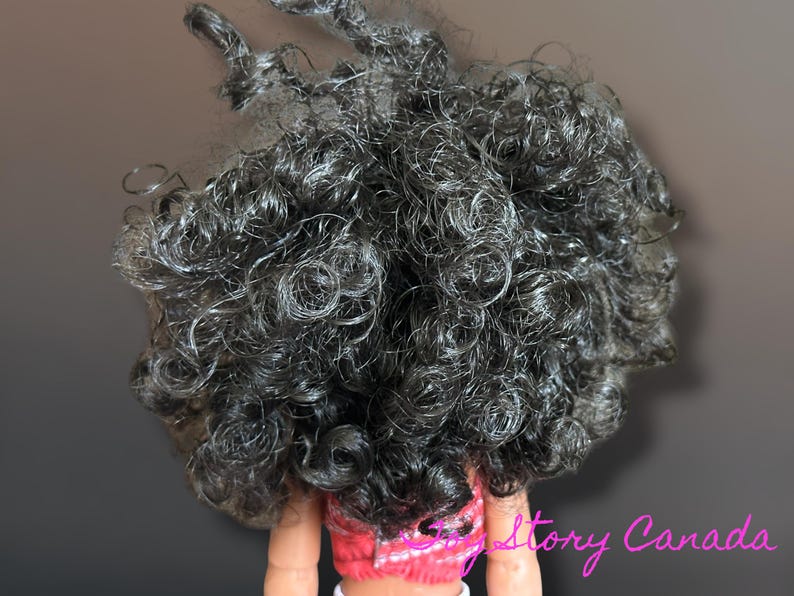 May include: Close-up of a doll's head with dark, tightly curled hair. The hair is a mix of black and gray, with individual curls and ringlets. The doll is wearing a pink and white top.