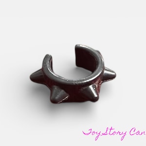 Può includere: Un ear cuff grigio scuro con un design a punte. Il gioiello a forma di U ha tre punte su ogni lato. Il gioiello è realizzato in materiale metallico ed è posto su uno sfondo bianco.