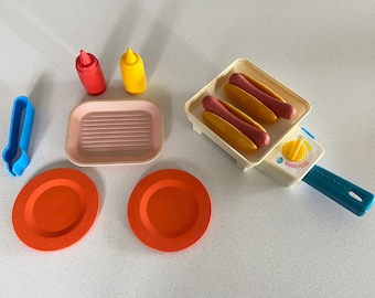 Puede incluir: Un juego de parrilla de hot dogs de juguete con dos hot dogs, una espátula, dos botellas de ketchup y mostaza, dos platos naranjas y una sartén de parrilla rosa.
