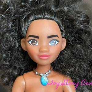May include: A doll with dark skin, brown eyes, and black curly hair. The doll has a pink mouth and a blue pendant necklace. The text "JoyStory Canada" is visible in the bottom right corner.
