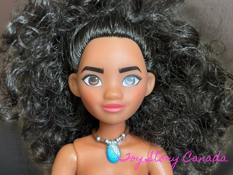 May include: A doll with dark skin, brown eyes, and black curly hair. The doll has a pink mouth and a blue pendant necklace. The text "JoyStory Canada" is visible in the bottom right corner.