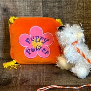 Peut inclure: Un jouet en peluche blanc avec une laisse rayée orange et blanche. Le jouet est assis à côté d'un petit coussin orange avec une fleur rose brodée dessus. La fleur a les mots "Puppy Power" brodés en violet.