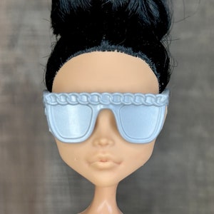 May include: Silver sunglasses with a chain detail on the frame. The sunglasses are on a doll with black hair.